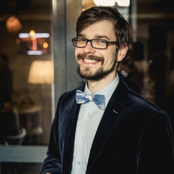 Mężczyzna w okularach i marynarce z muszką uśmiecha się do kamery w eleganckim otoczeniu.