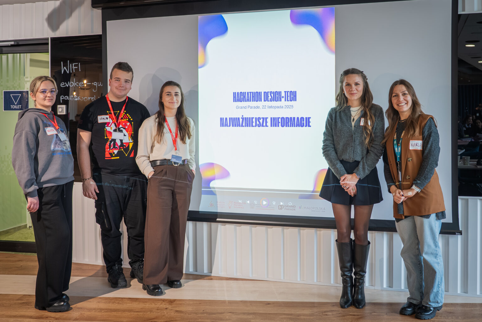 Pięciu uczestników hackathonu Design-Tech stoi przed ekranem z logiem wydarzenia, Grand Parade 22 listopada 2025.