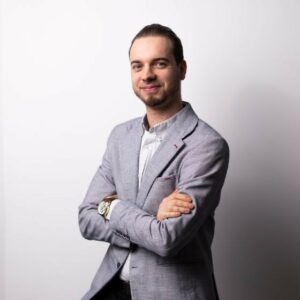 Portret profesjonalnego mężczyzny w szarej marynarce i białej koszuli, stojącego z założonymi rękami na szarym tle.