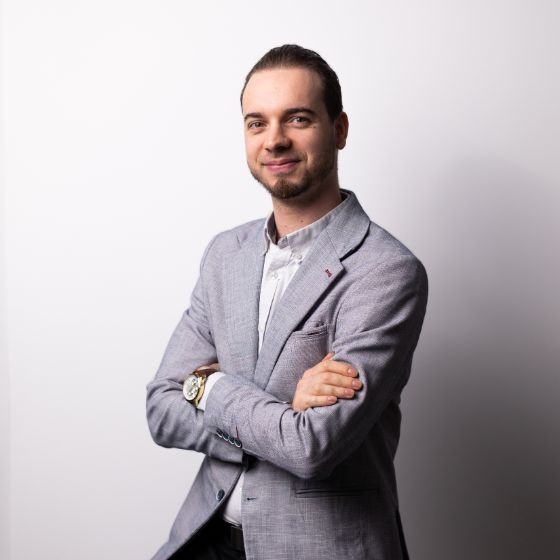 Portret profesjonalnego mężczyzny w szarej marynarce i białej koszuli, stojącego z założonymi rękami na szarym tle.