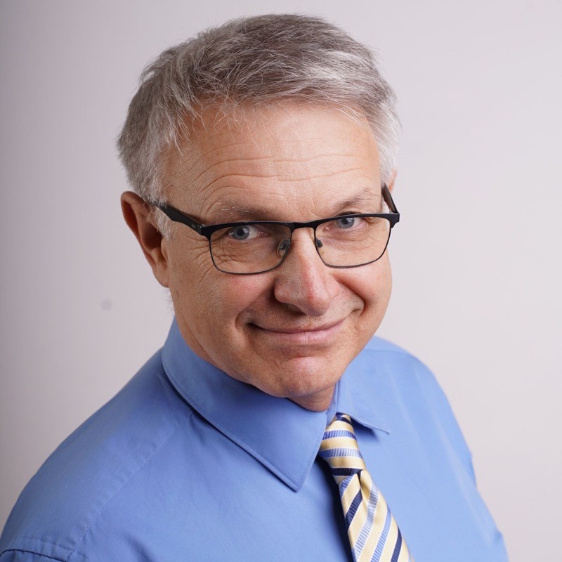 Portret profesjonalnego mężczyzny w niebieskiej koszuli i krawacie w paski na szarym tle.