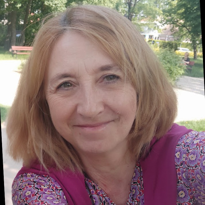 Kobieta w średnim wieku z brązowymi włosami, ubrana w fioletową bluzkę, uśmiechająca się do aparatu w ogrodzie.