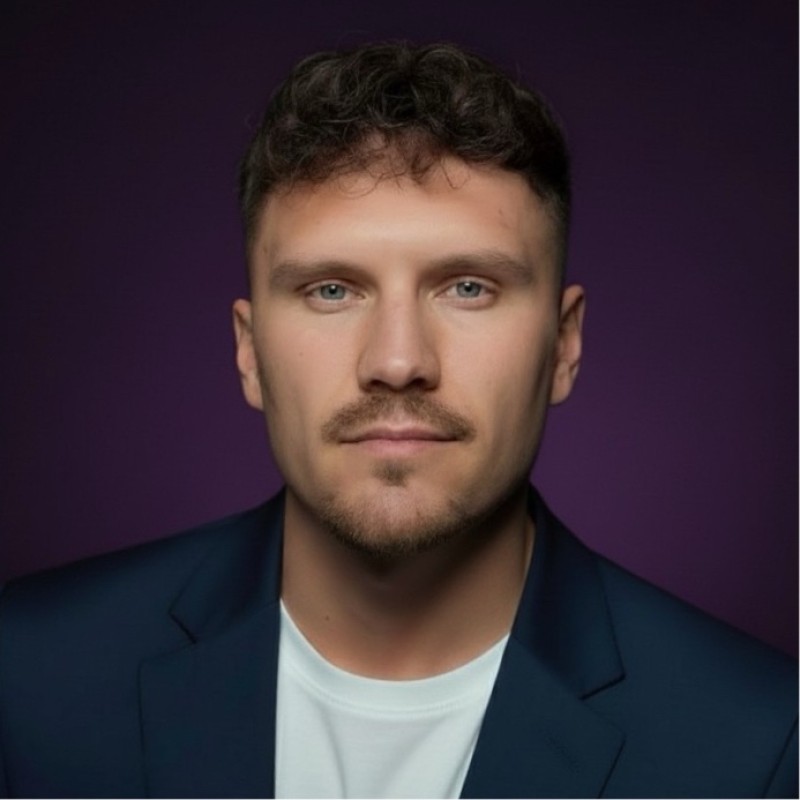 Portret mężczyzny z brązowymi włosami i zarostem w czarnej marynarce na fioletowym tle.