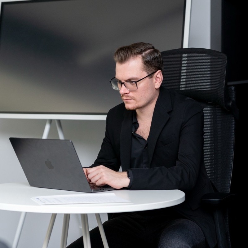 Mężczyzna w czarnej marynarce pracuje na laptopie przy białym stole w nowoczesnym biurze z monitorem