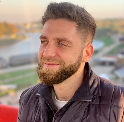 Mężczyzna z brodą i włosami upiętymi do góry, uśmiechający się do kamery na tle panoramy miasta.