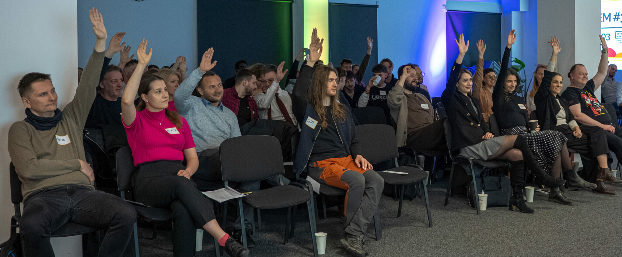 Grupa uczestników konferencji z podniesionymi rękami w nowoczesnej sali z niebieskim oświetleniem i projekcją na ekranie.