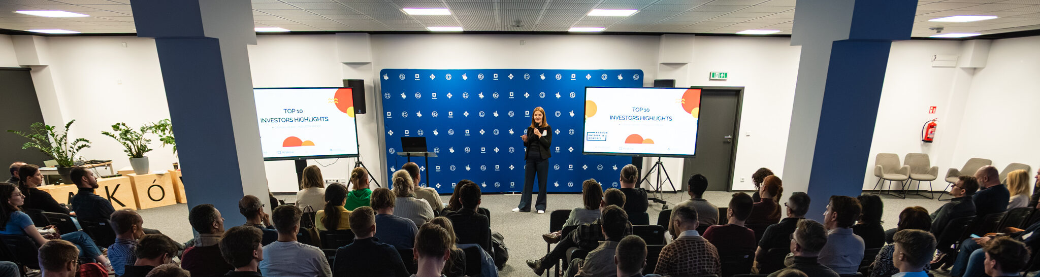 Konferencja biznesowa z prelegentem na scenie, publiczność siedzi na krzesłach, duże ekrany, niebieska ściana tła.