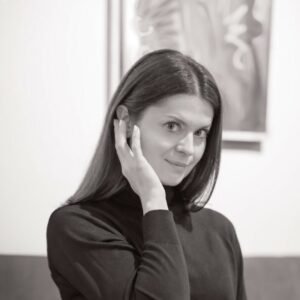 Portret kobiety z długimi czarnymi włosami, ręką przy twarzy, w czarnej bluzce, czarno-białe zdjęcie.